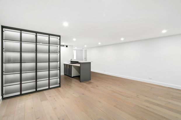 Bright, minimalist open-plan interior with pale timber flooring, white walls and ceiling, recessed downlights, a compact dark kitchen island, and a large black-framed glass-fronted built-in shelving or display unit along the left wall.