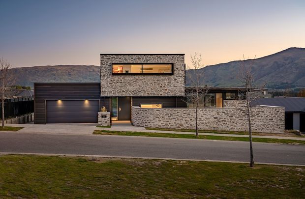 A contemporary multi-level residential facade at dusk, composed of grey stacked-stone walls, dark metal roofing and garage doors, and large horizontal glazed openings. A low stone boundary wall fronts the property, with a mountain landscape visible behind it.