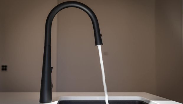Close-up product-style image of a tall matte black gooseneck kitchen tap with water running into a partially visible sink. The tap is set against a warm taupe wall and a light benchtop, with minimal surrounding context.