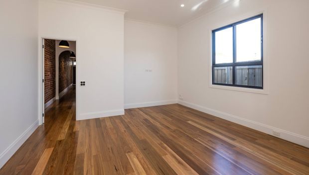 Empty, bright rectangular room with polished timber flooring, white painted walls and traditional white ceiling cornices and skirting boards. A large blue-framed window overlooks a fenced exterior, while an open doorway leads to a brick-lined hallway with timber floors and arched detailing. Recessed ceiling lights provide additional illumination.