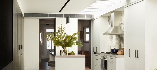 Bright contemporary kitchen viewed from the main living area, with a central white island, white cabinetry, integrated appliances, a stainless-steel rangehood, marble-look splashback, dark timber-framed doors, and a linear skylight or clerestory above.