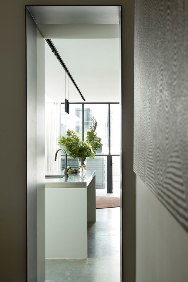 A narrow interior view through a dark-framed full-height mirror or opening into a bright contemporary kitchen. The scene shows a white island with an integrated sink and curved black tap, a pale stone or concrete worktop, pendant lighting, floor-to-ceiling glazing, and abundant greenery in a vase. The surrounding walls are neutral, with a textured monochrome wall surface visible on the right.