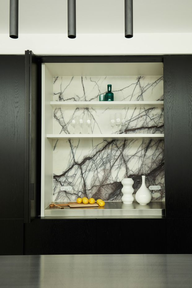 A close-up architectural interior detail showing a built-in black timber display cabinet with open shelving. The shelves and rear wall are clad in dramatic white marble with bold dark veining, displaying glassware, a green bottle, ceramic vases, and lemons. Black cabinetry extends across the wall, with cylindrical black ceiling lights above and a grey stone or concrete-like surface in the foreground. The composition is refined, dark, and highly material-focused.