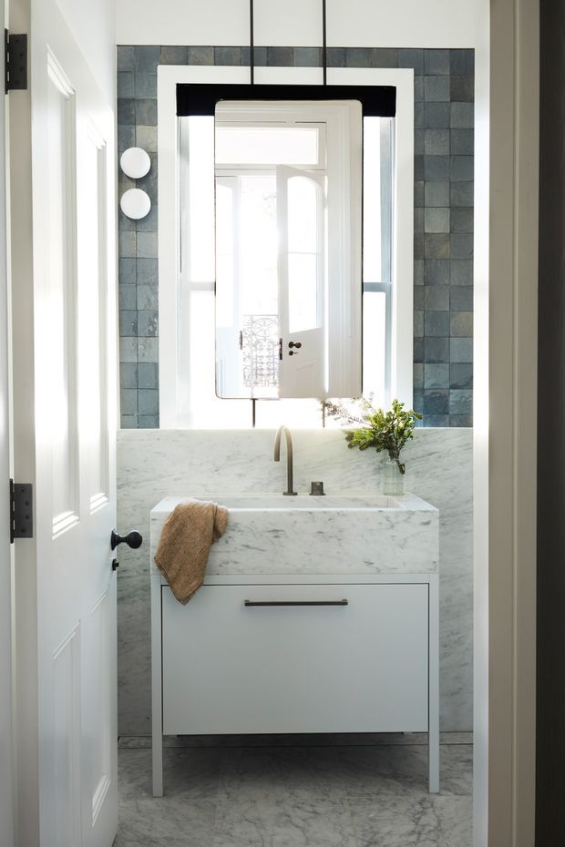 A compact, bright bathroom viewed through an open doorway. The room features a white floating-style vanity with a marble countertop and integrated backsplash, a single basin with brushed metal tapware, a large rectangular mirror, and dark blue-grey square wall tiles. White trim and a partially visible white door contrast with the tiled surfaces, while natural light enters from the window behind the mirror.