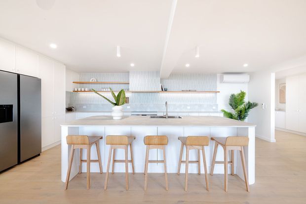 Bright contemporary open-plan kitchen dominated by a long white island with a pale stone benchtop and five timber bar stools. The room features white full-height cabinetry, a large dark refrigerator, pale timber flooring, a light blue-grey vertically textured tile splashback, open timber shelving, recessed ceiling lights, and indoor plants. A dining or living area is partially visible beyond the kitchen.