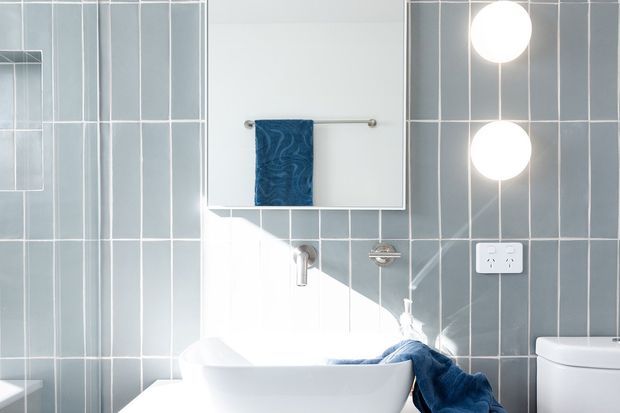 Bright contemporary bathroom with a freestanding white bathtub, white toilet, wall-mounted fixtures, mirrored cabinet, blue towel, and pale blue-grey vertically arranged wall tiles. Round wall lights and a white electrical outlet are visible on the tiled wall.