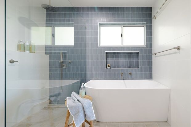 Bright contemporary bathroom with a freestanding white bathtub, a glass-enclosed shower, wall-mounted shower fixtures, and a recessed niche. The feature wall is clad in narrow vertical blue-grey tiles, complemented by pale floor tiles, white walls, chrome fittings, and two small horizontal windows. A timber stool with folded towels sits beside the bath.