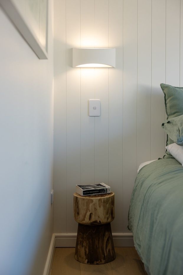 A close-up view of a calm bedroom corner with pale vertical wall panelling, a softly glowing modern wall sconce, and a bed dressed in muted sage-green bedding. A sculptural natural wood stump-style side table holds a book beside the bed. The space is softly lit with a restrained, natural palette and simple detailing.