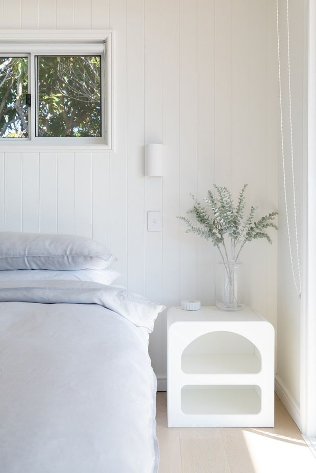 Bright, serene bedroom corner with a neatly made bed, white vertical wall panelling, a small framed window overlooking foliage, a simple wall light, and a rounded white bedside table styled with a glass vase of eucalyptus. Pale timber flooring and abundant natural light create a clean, airy feel.
