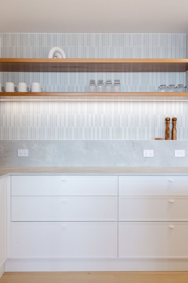 A bright, close-up view of a contemporary kitchen storage wall featuring white lower drawers and cabinetry, open timber shelves, pale blue-grey vertical tiled splashback, and a light grey stone or concrete-look benchtop and backsplash. White mugs and glassware are arranged on the shelves, with timber pepper mills adding a small warm accent. Light timber flooring is visible at the base.