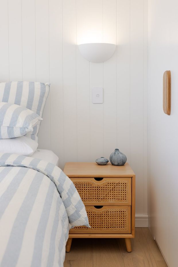 A softly lit bedroom vignette featuring a white vertical-panelled wall, a partially visible bed dressed in pale blue and white striped bedding, and a compact light timber bedside cabinet with woven cane drawer fronts. A simple wall sconce, small decorative vessels, and a wall-mounted timber detail complete the calm, coastal-inspired setting.