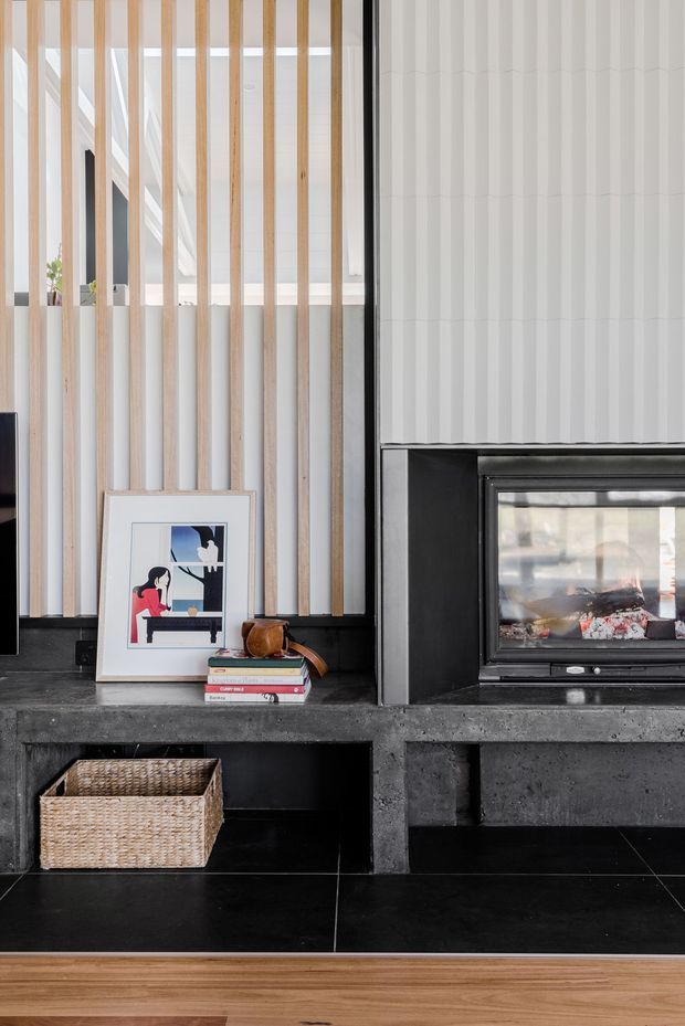 A close-up interior view of a contemporary living room fireplace wall. A black built-in fireplace and dark concrete hearth sit beneath a white textured wall finish, with vertical light timber battens screening glazing or an opening on the left. A framed artwork, books, and woven basket add restrained decorative detail. Black tiled flooring transitions to warm timber flooring in the foreground.