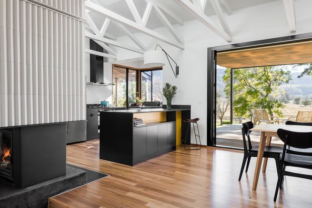 Bright open-plan kitchen and dining area with a black central island, timber flooring, white exposed roof trusses, vertical wall panelling, and a dark fireplace at left. Full-height glazing and an open sliding door connect the dining area to an outdoor deck and landscaped view. A compact dining table and chairs occupy the foreground.