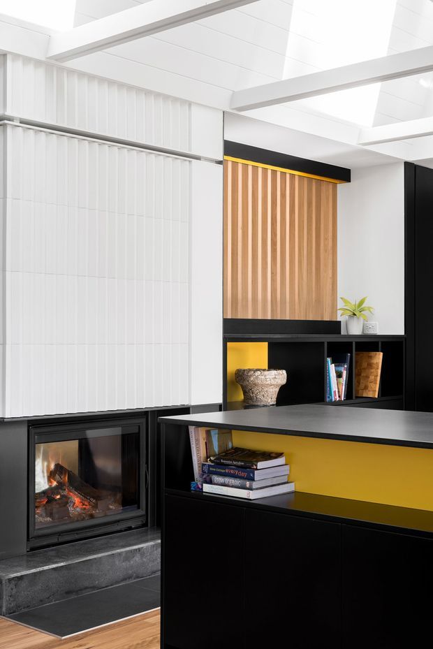 A bright contemporary interior featuring a black built-in fireplace, extensive black cabinetry and shelving, warm vertical timber wall panelling, and a vivid yellow accent recess. White ribbed wall panels and a high white ceiling with exposed linear beams create a crisp, graphic composition. Books, a planter, and a sculptural object are displayed in the joinery.