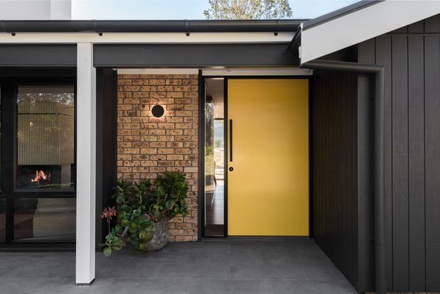Contemporary residential entrance beneath a projecting flat roof, featuring a bold mustard-yellow front door, dark vertical cladding, exposed warm brickwork, charcoal tiled paving, and a glazed side panel. A glimpse of a fireplace is visible through the adjacent window.
