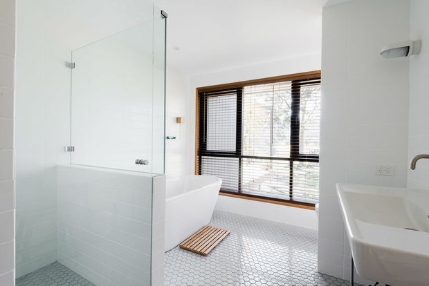 Bright contemporary bathroom with a freestanding white bathtub beside a large window, a glass-enclosed shower, wall-mounted double vanity, and pale hexagonal floor tiles. Dark timber-framed window blinds provide contrast against the predominantly white finishes.