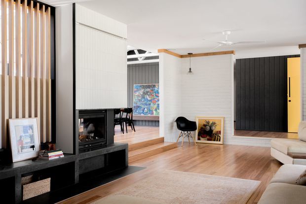 A bright contemporary open-plan living room with warm timber flooring, a large beige rug, and a partial cream sectional sofa in the foreground. A black built-in media and fireplace wall anchors the room, while vertical timber slats screen the entry or adjacent area. The space includes a raised dining zone with a table and chairs, a white painted brick feature wall, black paneled cabinetry or doors, exposed timber ceiling trim, and a ceiling fan. Colourful artwork and framed prints add visual accents.