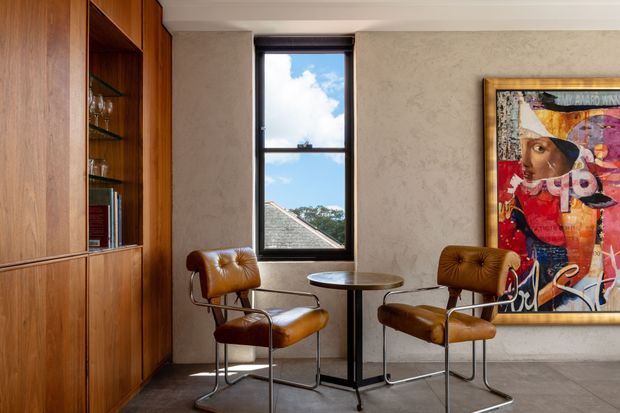 A refined interior sitting nook with two cognac leather lounge chairs and a small round pedestal table beside a tall black-framed window. Full-height warm timber built-in cabinetry with open glass shelving lines the left wall, while a textured plaster wall and colourful framed artwork create a sophisticated focal point. The room has a restrained, warm palette and abundant natural light.