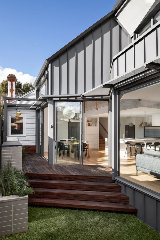 Bright exterior view of a contemporary multi-level residential extension with dark grey vertical cladding, extensive aluminium-framed glazing and large sliding glass doors opening onto a dark timber deck and stepped garden terrace. Through the glazing, a dining area and living room are visible, with a white kitchen, island seating and modern pendant lights. A small adjoining grey-clad structure and a brick chimney appear at the left.