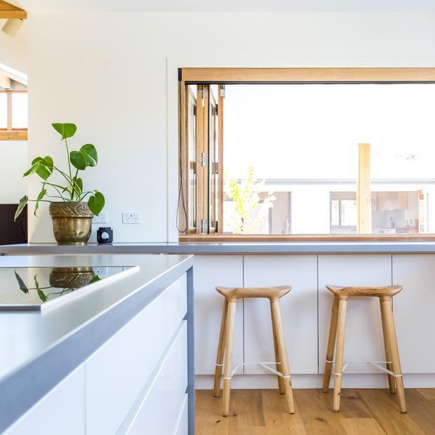 Bright contemporary kitchen with white cabinetry, a large reflective island, timber flooring, and two sculptural timber stools at a breakfast counter. A wide timber-framed servery window opens to an adjacent space, while a potted plant adds greenery.