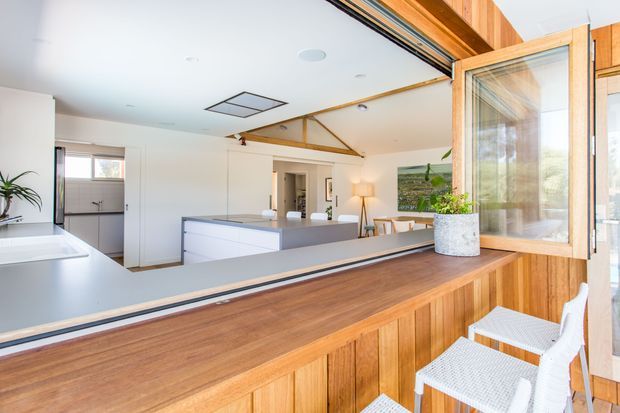 Bright open-plan kitchen and dining interior viewed from a timber-clad servery or bar counter. The space features white cabinetry, a pale benchtop, timber-framed folding windows, exposed timber ceiling structure, integrated downlights, indoor plants, and white bar stools. A large opening connects the kitchen to the living or dining area, creating an airy, indoor-outdoor entertaining feel.