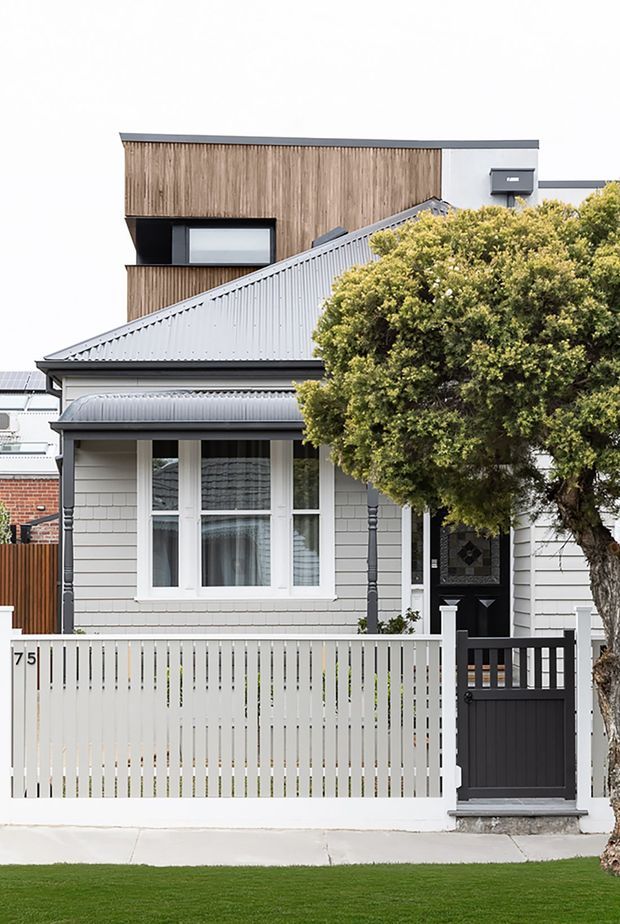 Street-facing exterior of a renovated heritage-style cottage with a white weatherboard facade, decorative front porch, pitched corrugated metal roof, white picket fence and dark entry gate. A contemporary upper-level addition is visible behind the original dwelling, clad in vertical timber boards with a simple flat roof and projecting dark-framed window. A mature tree partially obscures the right side.