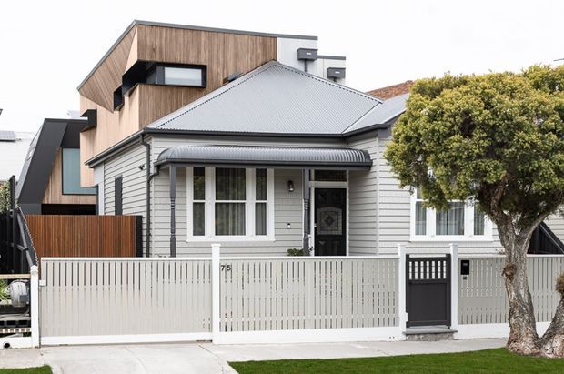 Street-facing view of a compact residential home combining a traditional single-storey weatherboard cottage frontage with a taller contemporary timber-clad addition behind it. The facade includes a pitched corrugated metal roof, white horizontal cladding, projecting windows, a covered entry, white picket-style fencing, and a dark pedestrian gate. A mature street tree partially screens the right side.
