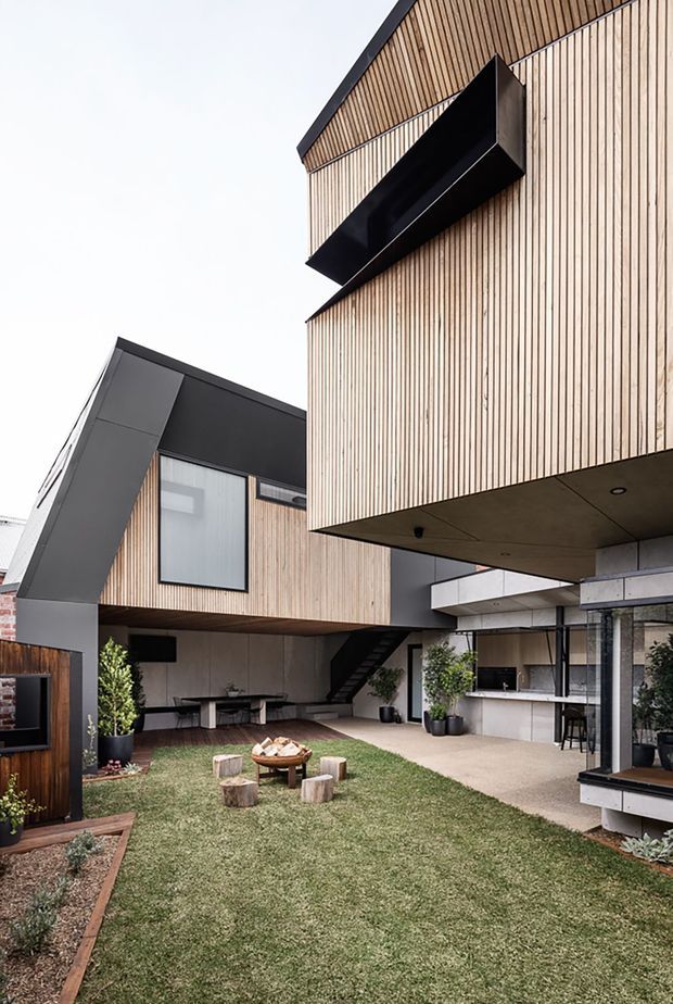 A contemporary multi-level residential courtyard viewed from outdoors, featuring projecting box-like volumes clad in fine vertical timber battens, dark charcoal metal-framed sections, and a broad cantilevered upper storey. The sheltered courtyard includes lawn, timber seating, potted plants, an outdoor dining area, stairs, and a partially visible outdoor kitchen or bar beneath the building. The composition highlights strong geometric forms, open-air circulation, and warm timber contrasted with dark cladding and concrete soffits.