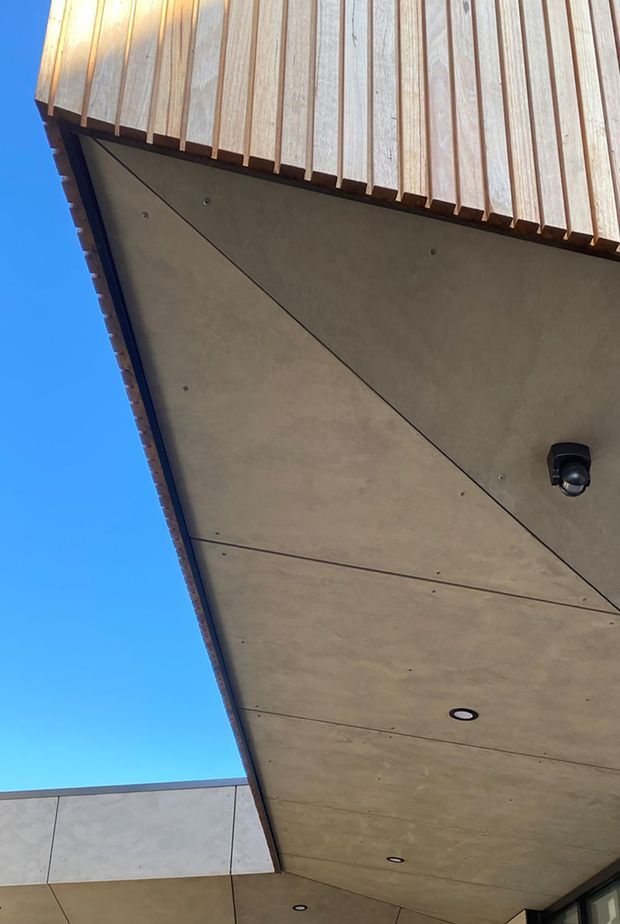 Upward-looking exterior architectural detail showing a broad soffit beneath a projecting roof or canopy. The soffit is formed from large smooth cement-look panels with visible joints, recessed downlights, and a black security camera. Above it is a warm timber-lined fascia or roof edge with closely spaced vertical battens. A pale panel-clad wall and blue sky are visible at the edges.