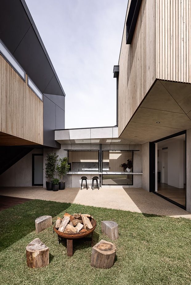 Contemporary multi-level courtyard house organised around a small grassed courtyard. The building combines vertical timber cladding, dark metal fascia and framing, and exposed concrete surfaces, with an outdoor bar or kitchen opening onto the courtyard. Large glazed openings connect the interior to the exterior, while a freestanding fire bowl and timber stump seats occupy the foreground.