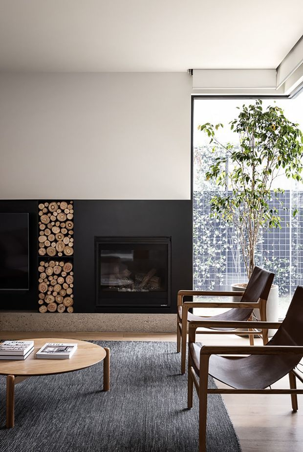 A refined contemporary living room with a black feature wall incorporating a built-in fireplace and a vertical display of stacked timber logs. A television is partially visible to the left. Large floor-to-ceiling glazing on the right brings in daylight and overlooks a planted courtyard or screened outdoor area. Mid-century-inspired timber lounge chairs, a round timber coffee table, and a textured grey rug furnish the room. The palette is restrained, combining warm timber, black, white, grey, and natural greenery.