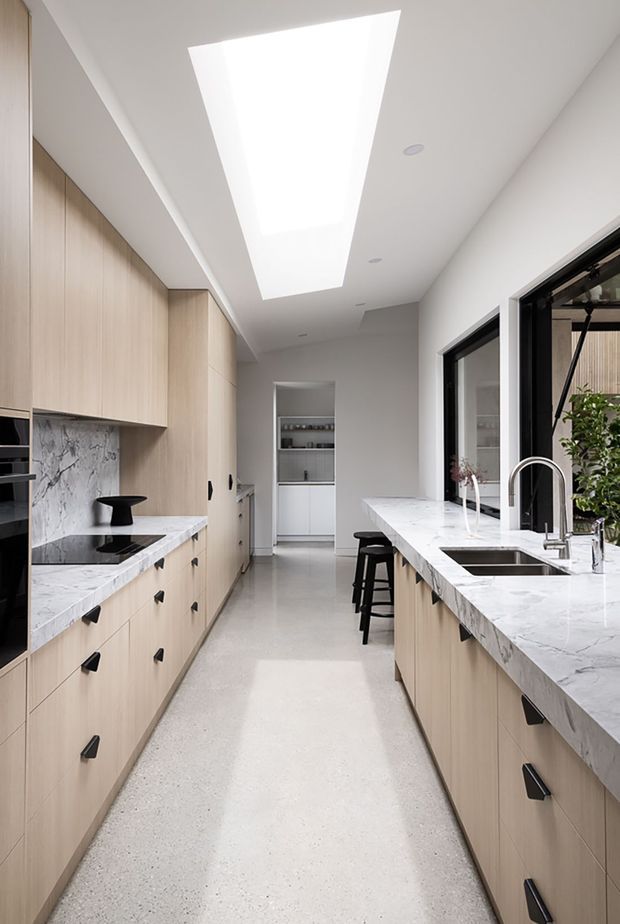 Bright contemporary galley kitchen with full-height light timber cabinetry on both sides, pale veined stone benchtops and splashback, black hardware, integrated appliances, a central walkway, and a large island with an undermount sink and tall chrome tap. A long ceiling skylight provides abundant natural light, while black-framed windows and a small pantry or shelving area are visible at the rear.