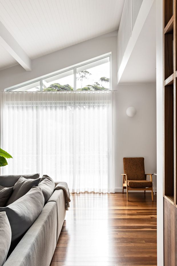 Bright, airy living room with a large grey sectional sofa, warm timber flooring, sheer full-height curtains, and a tall sloping white ceiling with exposed structural beams. A brown upholstered armchair sits beside the window, while built-in dark timber shelving frames the right side. High clerestory-style glazing brings in abundant natural light.