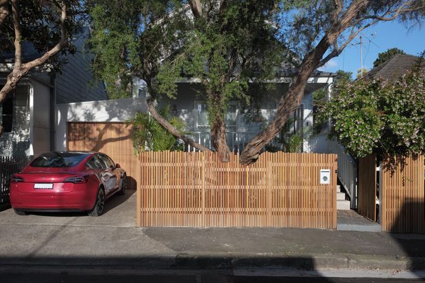 Street-facing view of a contemporary residential property with a timber slatted front fence and matching pedestrian gate, a concrete driveway, mature trees, and a partially visible dark-clad house behind the fence. A red car is parked in the driveway.