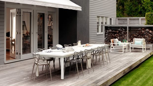 Contemporary outdoor dining and lounge area on a timber deck beside a grey weatherboard-clad house. A long white dining table with modern chairs sits beneath a retractable awning, with white-framed lounge seating and a built-in firewood storage feature along the rear wall. Folding glazed doors connect the deck to the interior.