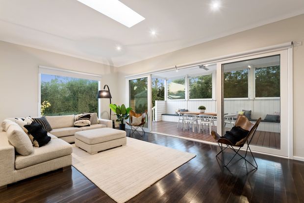 Bright, spacious living room with a large cream sectional sofa, ottoman, neutral area rug, black floor lamp, and leather sling chair. Dark polished timber flooring contrasts with light walls and ceiling. Wide glazed doors and windows open to a timber deck and enclosed outdoor area, with abundant greenery visible beyond.