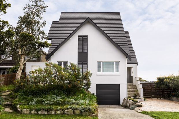 Front exterior view of a contemporary two-storey home with a steep, prominent gabled roof, white rendered walls, dark roof tiles, and dark-framed windows. The property includes an integrated single garage, paved driveway, landscaped garden beds, and a side stairway leading toward the entrance.