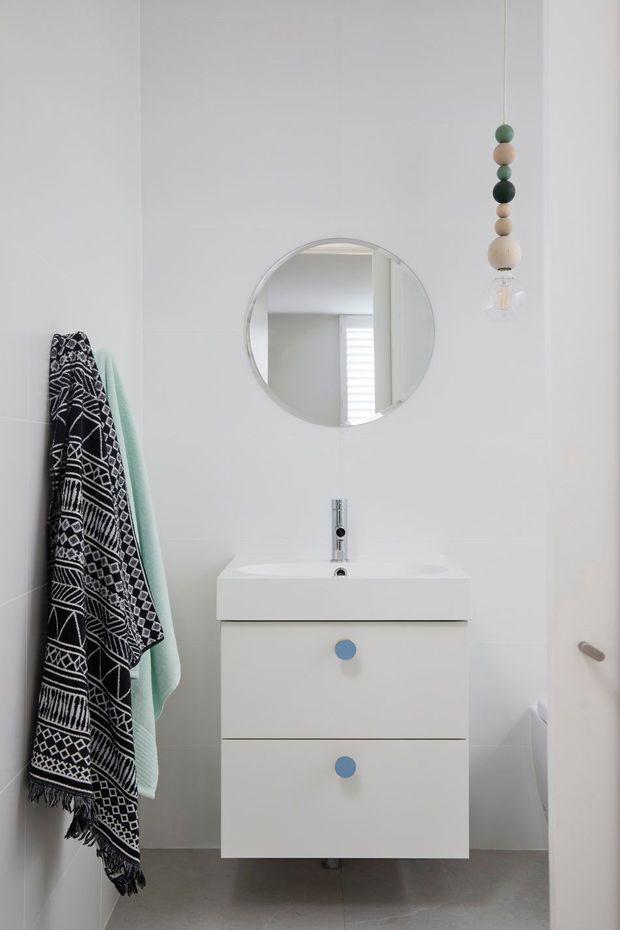 A compact, bright bathroom with white tiled walls, a wall-mounted white vanity with two drawers and blue circular pulls, a slim chrome basin tap, and a round wall mirror. A patterned black-and-white towel and pale green towel hang on the left, while a decorative pendant with wooden beads adds a playful detail. Grey floor tiles and a partially visible white door complete the space.