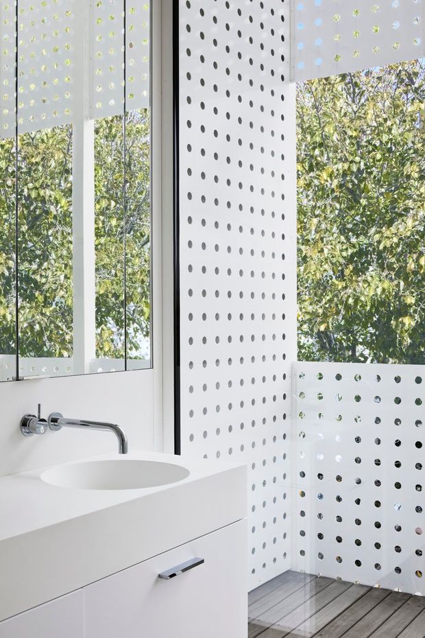 Bright contemporary bathroom with a minimalist white floating vanity, integrated oval basin, and wall-mounted chrome tapware. Floor-to-ceiling glazing and perforated white privacy screens create a dotted geometric pattern while allowing views to leafy greenery outside. The shower area appears enclosed by clear glass and has a grey timber-look floor.