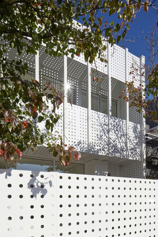 Exterior view of a contemporary multi-level building screened by perforated white facade panels and a matching perforated boundary wall. The panels create strong patterned shadows across the balconies and facade, with glazed openings and slender white structural framing visible behind. Framed by leafy branches against a clear blue sky.