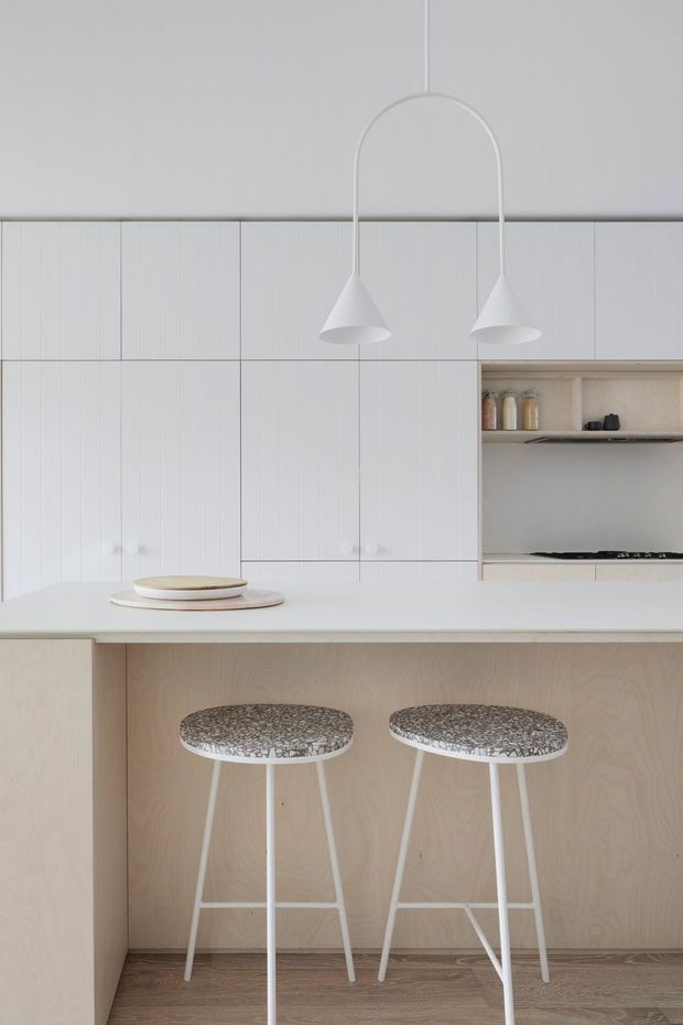 A bright, minimalist kitchen focused on a white island with a pale timber-clad breakfast bar and two stools. The kitchen has extensive white vertically panelled cabinetry, an integrated cooktop, open shelving with ceramics, and two sculptural white pendant lights. Pale timber flooring and restrained neutral finishes create a clean, calm atmosphere.