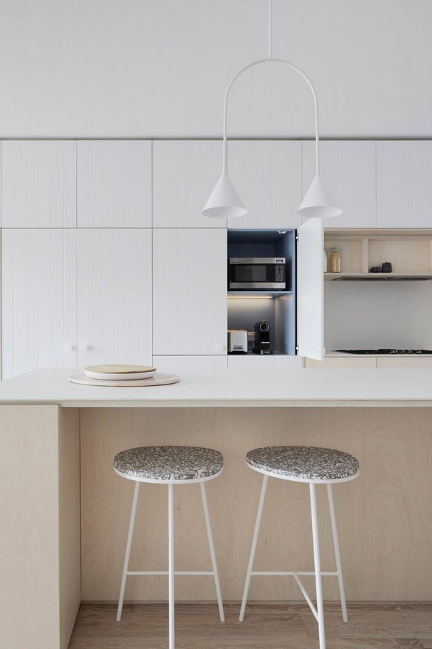 Bright minimalist kitchen with white vertically fluted cabinetry, integrated appliances, open shelving, a pale wood island front, and a light benchtop. Two sculptural white pendant lights hang over the island, which has two patterned bar stools. The restrained palette and clean lines create an airy, refined contemporary interior.