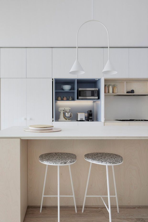 A bright, minimalist kitchen viewed straight on, featuring a long white island with two stools, pale timber or plywood cabinetry, white vertical-panel wall cabinetry, and an open blue-grey appliance niche containing a microwave and small appliances. A recessed open shelf displays jars and crockery. Two sculptural white pendant lights hang over the island, and the space has warm timber flooring with restrained neutral finishes.