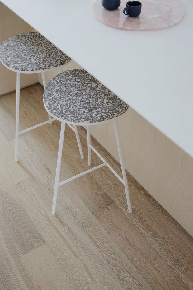 A close-up interior detail of a minimalist kitchen or breakfast bar, showing a white benchtop with pale timber cabinetry, two white-legged bar stools with grey terrazzo seats, and light timber flooring. A round pink stone tray with dark cups sits on the benchtop.