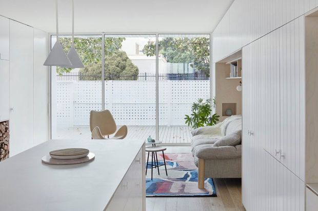 Bright open-plan contemporary living area viewed from the kitchen, with a large white island in the foreground, built-in white cabinetry along the right wall, a neutral sofa, sculptural pendant lights, timber flooring, and full-height glazing opening toward a small outdoor courtyard. The room has a clean, airy, minimalist feel with colourful patterned rug accents.