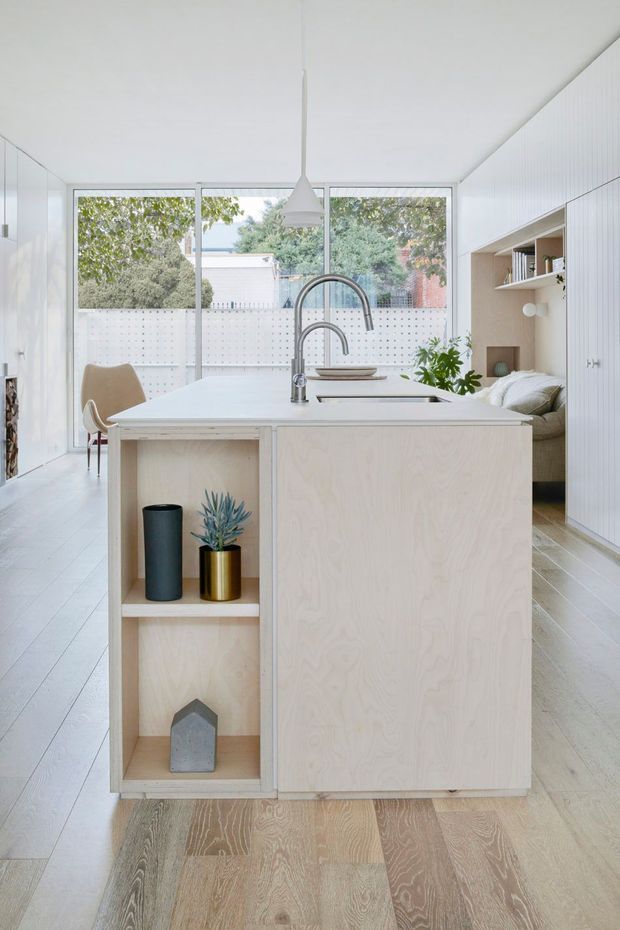Bright, compact open-plan kitchen with a pale plywood island featuring open display shelving, an integrated sink and tall curved chrome tap. White vertical-panel cabinetry lines the walls, with a built-in banquette and shelving at the rear. Pale timber flooring, white walls, a simple pendant light, and large glazed doors create an airy, minimalist interior connected to a small outdoor area.