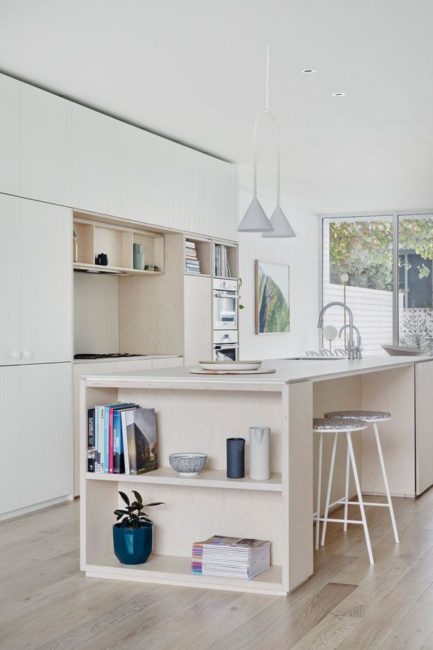 Bright, compact contemporary kitchen with a pale timber island featuring open display shelving, white integrated cabinetry, a light benchtop, gas cooktop, and double-bowl sink with tall arched tap. Two white pendant lights hang above the island, with bar stools tucked beneath the overhang. Light timber flooring, white vertical wall panelling, and a large window create an airy, Scandinavian-influenced interior.