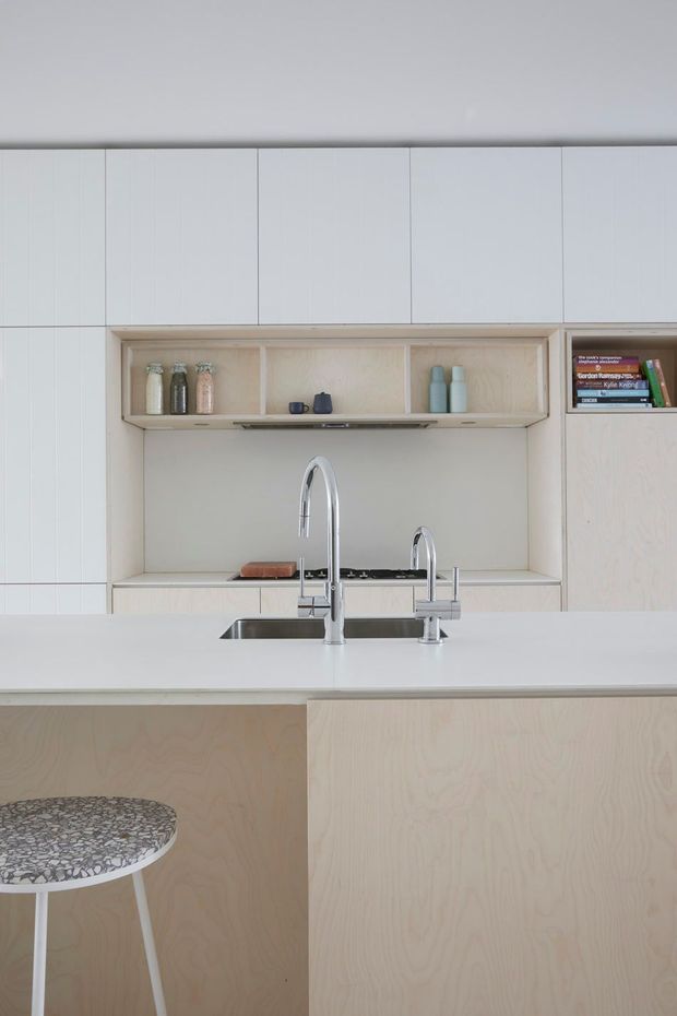 A minimalist contemporary kitchen viewed from the island. Pale timber plywood cabinetry and wall panelling surround a white island benchtop with integrated stainless-steel sinks and tall chrome taps. An open recessed shelf displays glass jars, cups, bottles, and a few books, while a patterned round stool is partially visible in the foreground.
