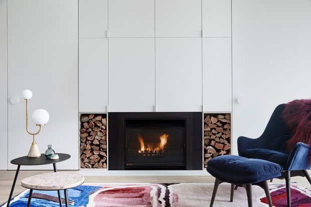 A refined contemporary living room centered on a black built-in fireplace with stacked firewood niches on both sides. Full-height white built-in cabinetry creates a clean wall composition, while a dark navy lounge chair, matching ottoman, black side table, sculptural lamp, and colourful abstract rug add contrast and visual interest.