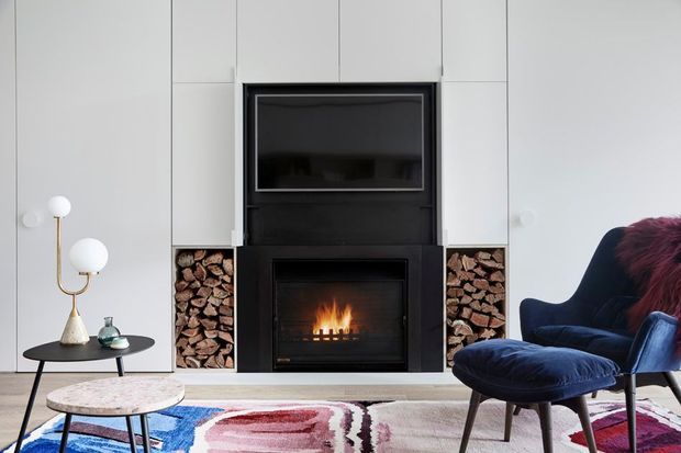 A refined contemporary living room centered on a black fireplace and television surround, with neatly stacked firewood niches on both sides. White full-height wall panelling creates a clean backdrop, while a dark blue lounge chair and ottoman, small round coffee tables, sculptural floor lamp, and colourful abstract rug add contrast. The fireplace is lit, giving the room a warm focal point.